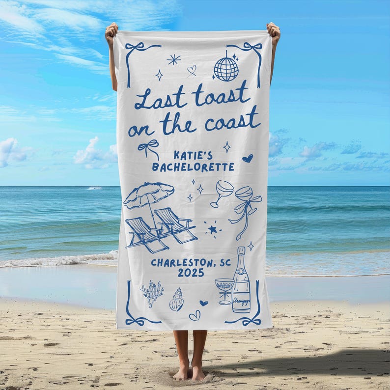 May include: White beach towel with blue text that reads "Last toast on the coast Katie's Bachelorette Charleston, SC 2025". The towel also features a blue illustration of a beach umbrella, two lounge chairs, champagne glasses, and a bottle of champagne.