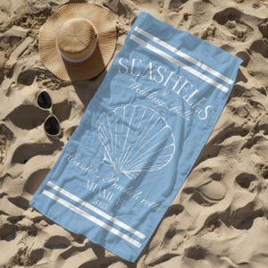 May include: A light blue beach towel with white text that reads "Seashells & Wedding Bells" and a white seashell graphic. The text also reads "Cassie's Bachelorette Miami, FL 2025".