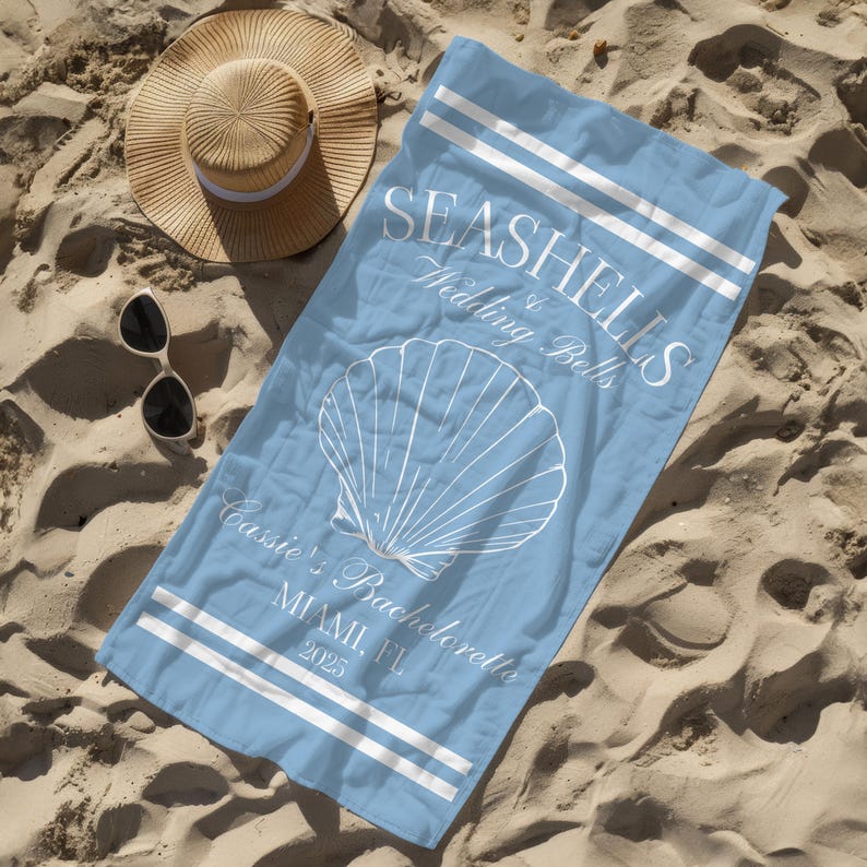 May include: A light blue beach towel with white text that reads "Seashells & Wedding Bells" and a white seashell graphic. The text also reads "Cassie's Bachelorette Miami, FL 2025".