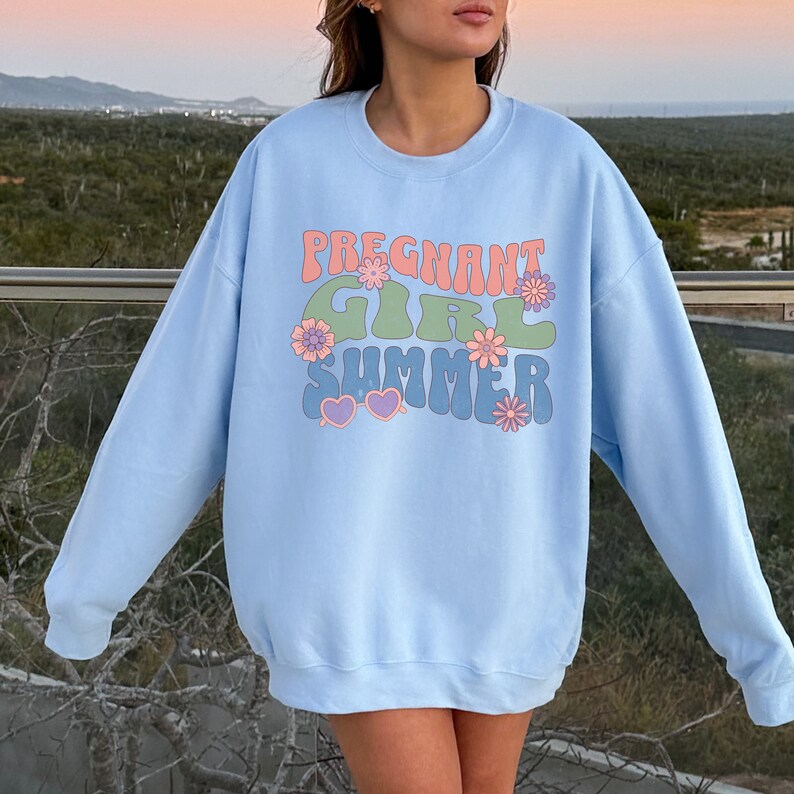 Puede incluir: Sudadera azul claro con un estampado gr&aacute;fico del texto "Pregnant Girl Summer" en una fuente retro con flores y corazones.