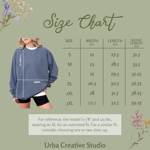 Op de afbeelding: Maattabel voor een blauwe sweatshirt, metingen in inches. De tabel bevat maten S tot 3XL, met bijbehorende breedte, lengte en mouwlengte metingen. Het model draagt een XL.