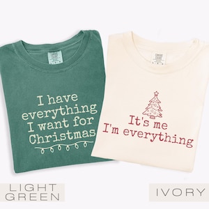 May include: Two t-shirts, one light green and one ivory, with festive text. The light green shirt says "I have everything I want for Christmas" with string lights. The ivory shirt says "It's me I'm everything" with a Christmas tree.
