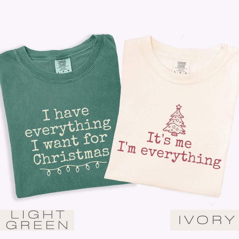 May include: Two t-shirts, one light green and one ivory, with festive text. The light green shirt says "I have everything I want for Christmas" with string lights. The ivory shirt says "It's me I'm everything" with a Christmas tree.