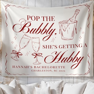 Puede incluir: Una pancarta de tela blanca con texto e ilustraciones en color burdeos. La pancarta dice "Pop the Bubbly, She's Getting a Hubby" con imágenes de copas de champán, una botella de champán en una cubitera y el texto "Hannah's Bachelorette Charleston, SC 2025".