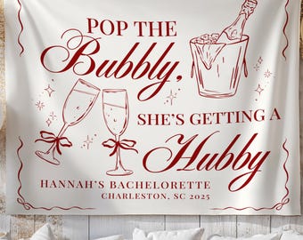 May include: A white fabric banner with burgundy text and illustrations. The banner reads "Pop the Bubbly, She's Getting a Hubby" with images of champagne glasses, a champagne bottle in an ice bucket, and the text "Hannah's Bachelorette Charleston, SC 2025".