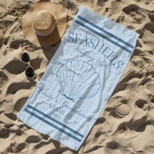 May include: A light blue beach towel with the text "Seashells & Wedding Bells" and a seashell graphic. The text "Cassie's Bachelorette Miami, FL. 2025" is also printed on the towel.