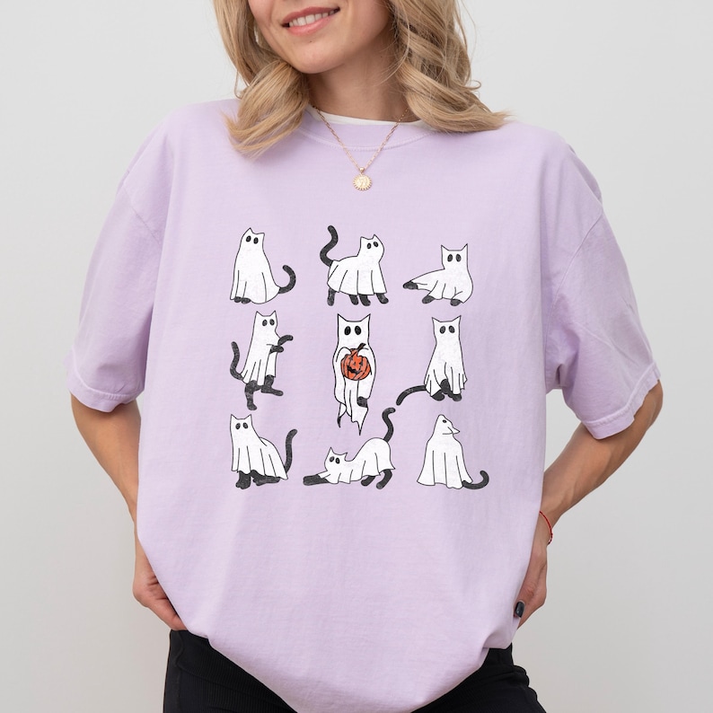 Puede incluir: Una camiseta lavanda con un gr&aacute;fico de nueve gatos blancos vestidos de fantasmas. Un gato sostiene una peque&ntilde;a calabaza naranja.