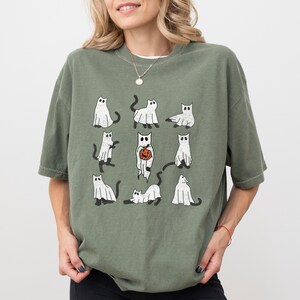 Puede incluir: Una camiseta verde con un gr&aacute;fico de gatos blancos vestidos de fantasmas. Algunos de los gatos sostienen calabazas. La camiseta tiene cuello redondo y mangas cortas.