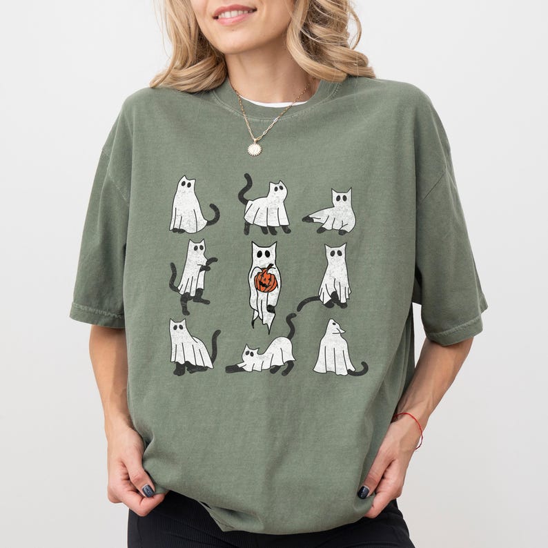 Puede incluir: Una camiseta verde con un gr&aacute;fico de gatos blancos vestidos de fantasmas. Algunos de los gatos sostienen calabazas. La camiseta tiene cuello redondo y mangas cortas.