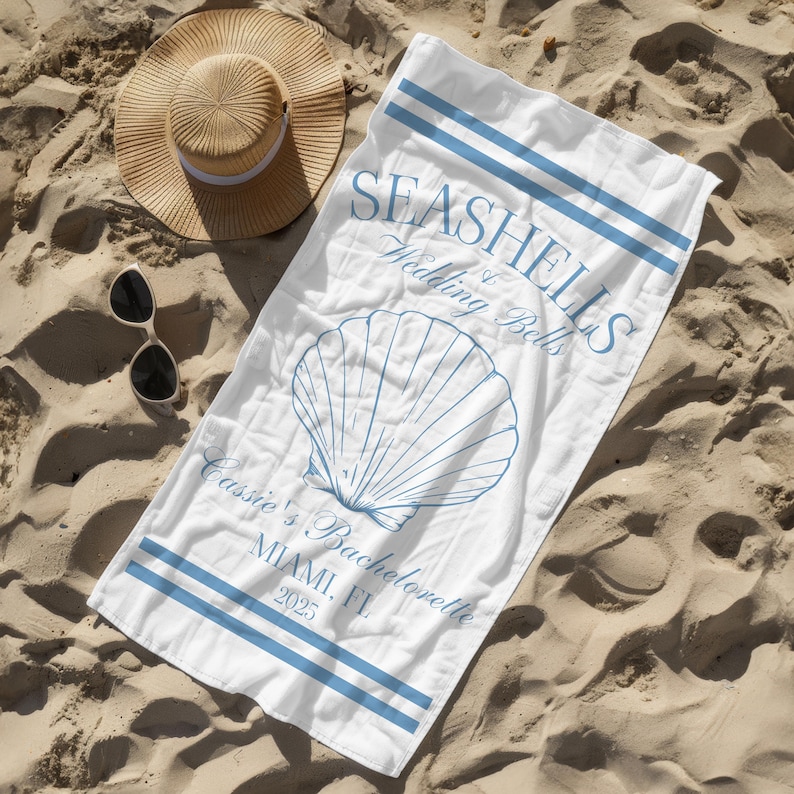 May include: White beach towel with blue stripes and the text "Seashells & Wedding Bells" and "Cassie's Bachelorette Miami, FL 2025". A seashell illustration is in the centre of the towel.