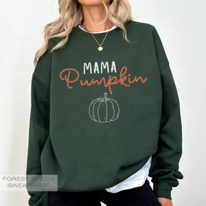 Pu&ograve; includere: Felpa verde bosco con il testo "Mama Pumpkin" in lettere bianche e arancioni. Un piccolo grafico di zucca si trova sotto il testo.