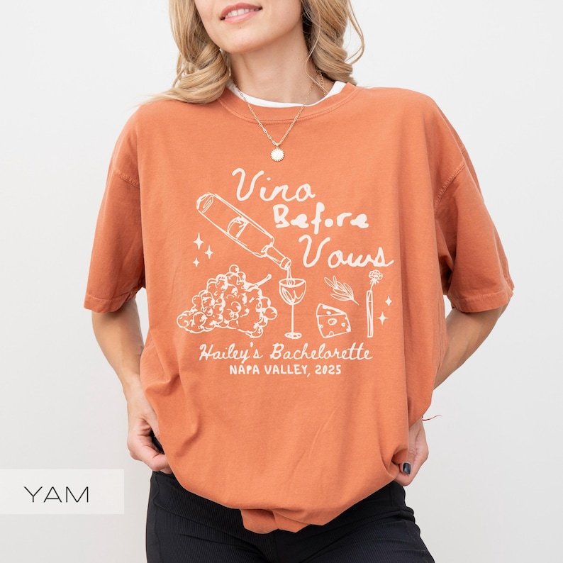 以下が含まれることがあります： 白の文字とグラフィックが入った、レンガ色のTシャツ。「Vino Before Vows」と書かれており、ワインボトル、ブドウ、ワイングラス、チーズ、花のイラストが描かれています。下には「Hailey's Bachelorette, Napa Valley, 2025」と書かれています。