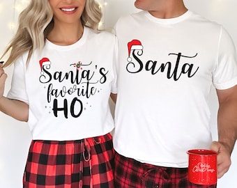 Può includere: Una coppia indossa magliette bianche abbinate con testo a tema natalizio. La maglietta della donna dice "Santa's favorite HO" con una grafica di cappello di Babbo Natale e bastoncino di zucchero. La maglietta dell'uomo dice "Santa" con una grafica di cappello di Babbo Natale. Entrambi indossano pantaloni da pigiama a quadri rossi e neri.