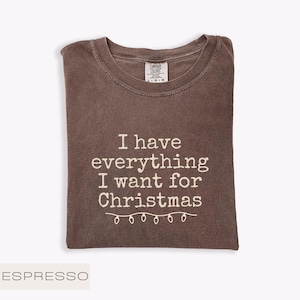 May include: An espresso-coloured t-shirt with the white text "I have everything I want for Christmas" and a string of lights graphic. The shirt is folded and has a Comfort Colors tag.