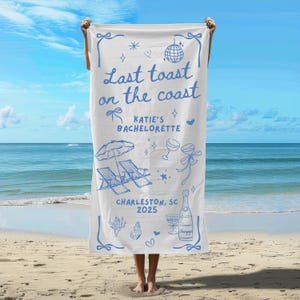 May include: White beach towel with blue text that says "Last toast on the coast Katie's Bachelorette Charleston, SC 2025". The towel also has a blue illustration of a beach umbrella, two lounge chairs, a bottle of champagne, and two champagne flutes.
