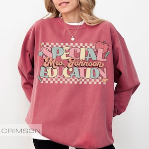Op de afbeelding: Kleur crimson sweatshirt met de tekst "SPECIAL Mrs. Johnson EDUCATION" in een retro lettertype. Het ontwerp bevat een appel, sterren en een geruite achtergrond. De sweatshirt is een crew neck stijl.