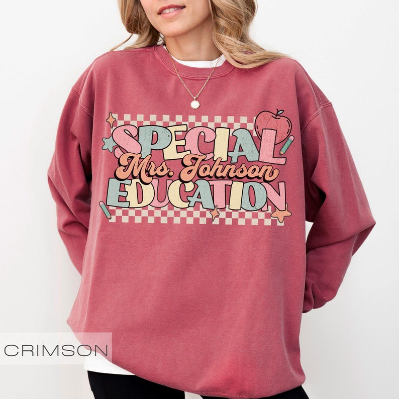 Op de afbeelding: Kleur crimson sweatshirt met de tekst "SPECIAL Mrs. Johnson EDUCATION" in een retro lettertype. Het ontwerp bevat een appel, sterren en een geruite achtergrond. De sweatshirt is een crew neck stijl.