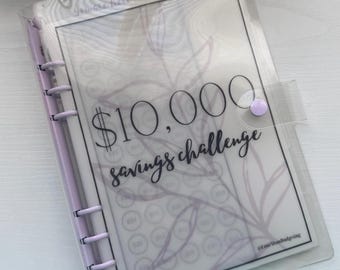 May include: A clear plastic A6 binder with a light purple spine and snap closure. The cover features a printed insert titled $10,000 savings challenge in black text, with a faint botanical line drawing and circular icons for tracking progress.