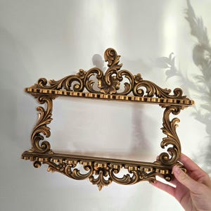 May include: Ornate gold-colored wall shelf with a rectangular opening. The shelf features intricate scrollwork, floral accents, and a detailed border. The design is symmetrical, with a central floral motif at the top. The shelf is held by a hand.