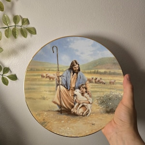 Puede incluir: Un plato decorativo redondo de cerámica con borde dorado que presenta una escena pintada de una figura barbuda con túnica color canela y chal azul sosteniendo un cayado de pastor y un cordero, con un niño y un rebaño de ovejas en un campo.