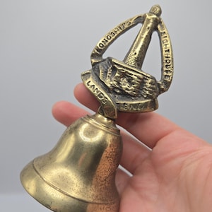 Può includere: Una campana in ottone con un manico decorativo a forma di faro. Il manico presenta il testo in rilievo "LONGSHIPS LIGHTHOUSE" e "LANDS END" alla base. La campana ha una finitura metallica liscia con segni visibili di età e patina.