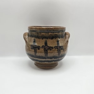Op de afbeelding: Een handgemaakte keramische pot in bruin en zwart. De pot heeft twee handvatten en een decoratief patroon van drie gestileerde figuren. De pot heeft een diameter van ongeveer 15 cm en is 13 cm hoog.