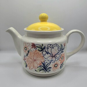 May include: Cream-coloured ceramic teapot with a yellow lid and handle. The teapot features floral patterns in peach, blue, and green. The lid has a yellow knob. A classic piece for serving tea.
