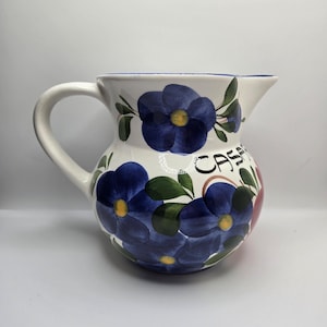 May include: White ceramic jug with a curved handle, decorated with large blue flowers, green leaves, and yellow centres. The word "CASA" is written on the side. The jug has a blue rim.