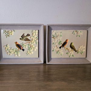 Puede incluir: Un par de placas de pared rectangulares con relieves tridimensionales de aves posadas en ramas florecientes. Cada placa está encerrada en un marco gris claro acanalado, con paneles centrales que muestran motivos florales de color crema.
