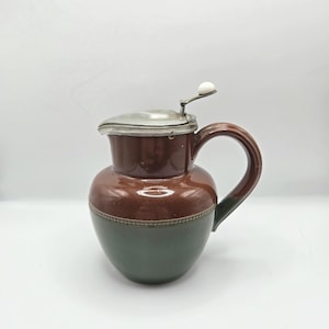 Peut inclure: Une cruche en céramique marron et verte avec une anse et un couvercle en métal. La cruche a une forme arrondie avec un dessus marron foncé et une base verte. Le couvercle a un petit bouton blanc. La cruche est probablement pour servir des boissons.