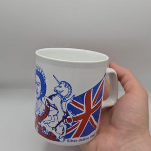 Peut inclure: Tasse en céramique blanche avec un motif bleu et rouge représentant le drapeau britannique, une licorne et un portrait de femme. La tasse porte l'inscription "Silver Jubilee 1952-1977". La tasse a une anse blanche.