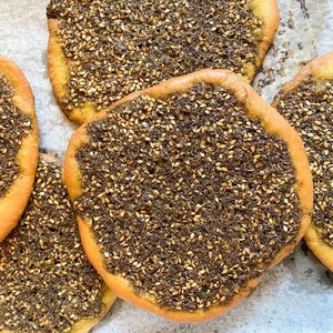 May include: Five round flatbreads topped with a mixture of sesame seeds and za'atar, a Middle Eastern spice blend, on a white parchment paper background.