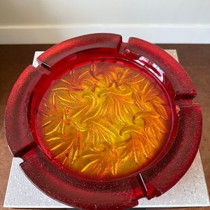 Może przedstawiać: Popielniczka ze szkła czerwonego z wytłoczonym wzorem kwiatowym w kolorze żółtym. Popielniczka ma szeroki, zaokrąglony brzeg i głębokie wgłębienie.