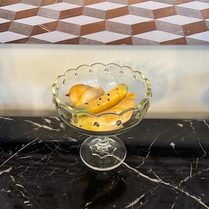 Könnte beinhalten: Eine klare Glasschüssel mit einem gewellten Rand und einem Stiel, gefüllt mit gelben und orangefarbenen Frucht-Schnitzereien. Die Schüssel steht auf einer schwarzen Marmorfläche.