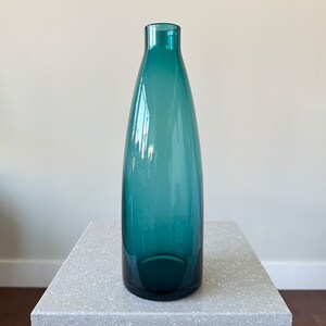 Peut inclure: Un vase en verre bleu turquoise avec un fond arrondi et un col étroit. Le vase est posé sur une surface grise mouchetée.