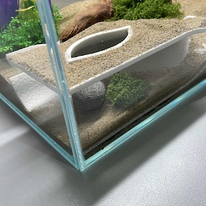 Peut inclure: Un terrarium en verre avec une base de sable blanc et une structure en plastique blanc avec un trou en haut. Le terrarium contient &eacute;galement un petit rocher gris, de la mousse verte et un morceau de bois flott&eacute;.