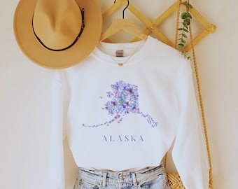 May include: White sweatshirt with a purple floral design of the state of Alaska and the word "ALASKA" printed below the design. The sweatshirt is hanging on a wooden hanger with a straw hat and a woven bag hanging nearby.