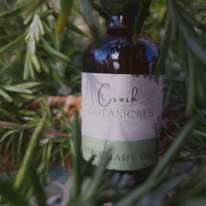 May include: A brown glass bottle with a dropper top and a white label that reads "Crush Botanicals Rosemary Oil". The bottle is partially obscured by green rosemary sprigs.