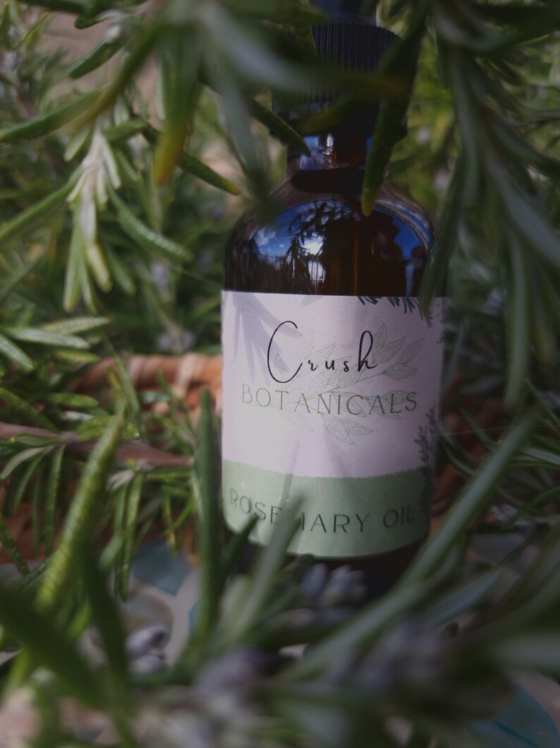 May include: A brown glass bottle with a dropper top and a white label that reads "Crush Botanicals Rosemary Oil". The bottle is partially obscured by green rosemary sprigs.