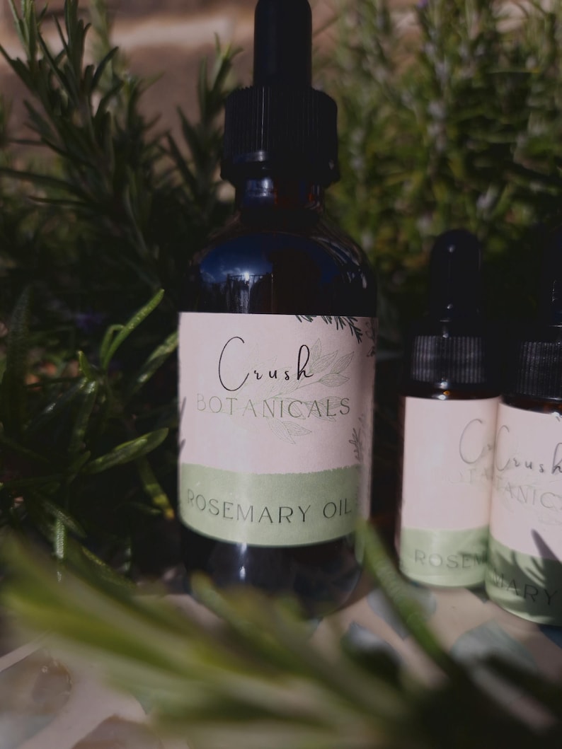 May include: Two glass bottles with droppers, one larger than the other, both filled with a dark liquid. The larger bottle has a white label with the text "Crush Botanicals Rosemary Oil". The smaller bottle has a white label with the text "Crush Botanicals Rosemary Oil". Both bottles are sitting on a green leafy surface.