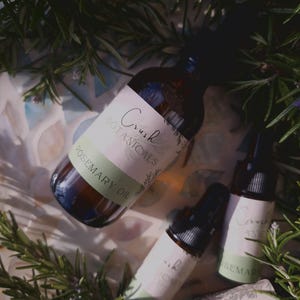 May include: Three glass bottles with dropper tops filled with rosemary oil. The bottles have white labels with green accents and the text "Crush Botanicals Rosemary Oil". The bottles are surrounded by green rosemary sprigs.