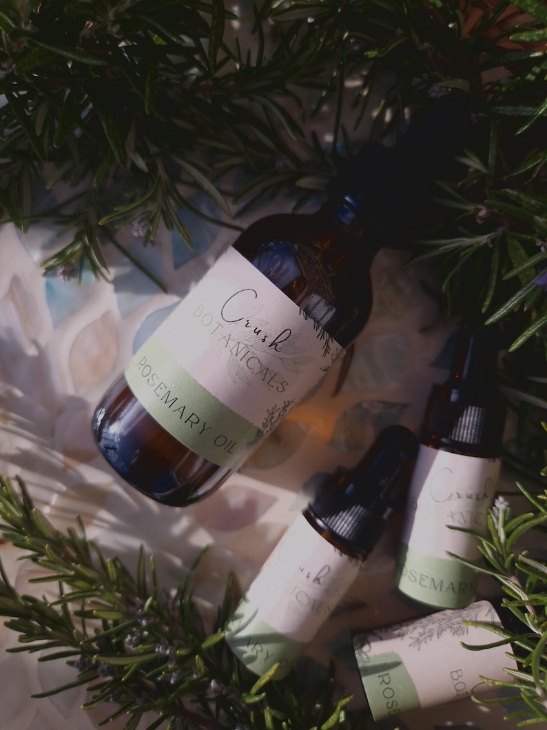 May include: Three glass bottles with dropper tops filled with rosemary oil. The bottles have white labels with green accents and the text "Crush Botanicals Rosemary Oil". The bottles are surrounded by green rosemary sprigs.
