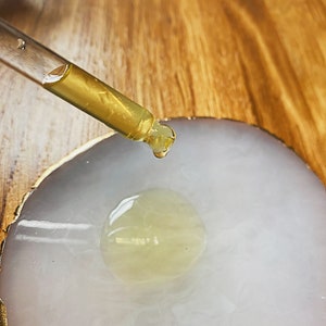 May include: A clear glass dropper with a yellow liquid is being used to drop a single drop of liquid onto a white, round, agate coaster with gold trim.
