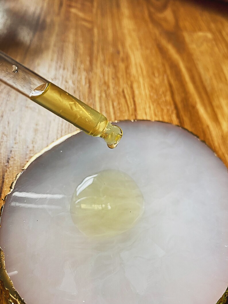 May include: A clear glass dropper with a yellow liquid is being used to drop a single drop of liquid onto a white, round, agate coaster with gold trim.