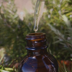 May include: A glass dropper dispenses liquid from a dark brown glass bottle. The bottle has a wide mouth and a screw-on lid. The background is blurred and shows green foliage.