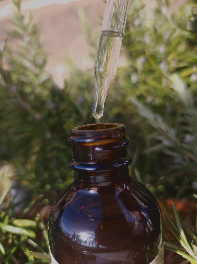 May include: A glass dropper dispenses liquid from a dark brown glass bottle. The bottle has a wide mouth and a screw-on lid. The background is blurred and shows green foliage.