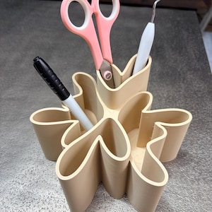 May include: A beige, flower-shaped desk tidy containing pink scissors, a black marker, and a white tool. The organiser has multiple compartments for storing pens, pencils, and other small office supplies. The design is modern and functional.