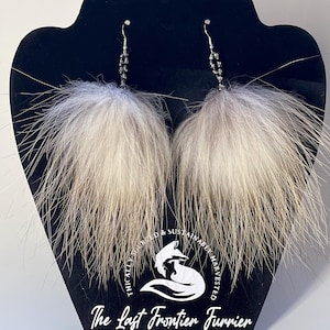 May include: A pair of fluffy, white and gray fur earrings with beaded accents. The earrings are displayed on a black stand with the text "The Last Frontier Furrier" and a fox logo. The earrings have a silver hook.