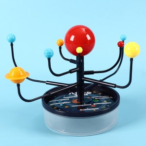 May include: A colorful solar system model with a red sun and eight planets on black arms. The base is black with a white background and a printed image of the solar system.