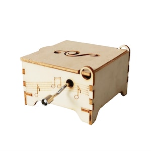 Może przedstawiać: Drewniana skrzynka muzyczna z wzorem klucza wiolinowego na górze. Skrzynka ma kluczyk do nakręcania i odtwarza muzykę po nakręceniu.